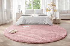 Amazon Basics fluffy washable round area rug in soft pink placed under a bed in a bright minimalist bedroom with wooden floors and natural light
